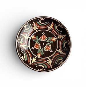 May include: A round, dark brown ceramic plate with a decorative floral design. The plate features a central floral motif in red, green, and yellow, surrounded by a repeating pattern of curved lines and floral elements in similar colors. The rim is a lighter brown.