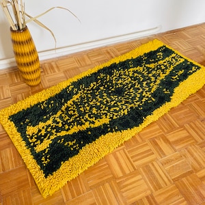 Op de afbeelding: Een geel en groen shag tapijt met een geometrisch patroon. Het tapijt is rechthoekig en heeft een gefranst randje.