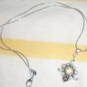 May include: Silver-toned necklace with a floral pendant. The pendant features a central yellow gemstone surrounded by smaller green, white, and brown stones. The necklace has a delicate chain and a lobster clasp.