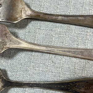 May include: Three silver-toned metal forks with visible tarnish and wear. The handles feature engraved text including Madison Silver Plate and National Silver Plate, along with a small triangular hallmark symbol on the top fork handle.