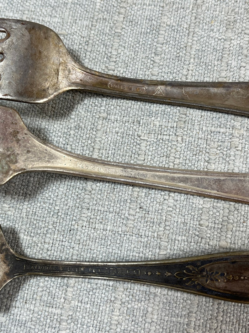 May include: Three silver-toned metal forks with visible tarnish and wear. The handles feature engraved text including Madison Silver Plate and National Silver Plate, along with a small triangular hallmark symbol on the top fork handle.
