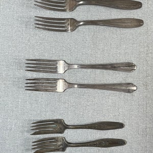 May include: A collection of six silver-toned metal forks arranged in three pairs. The top two forks have smooth handles, the middle two feature a fluted design, and the bottom two display an intricate embossed pattern on the handles.