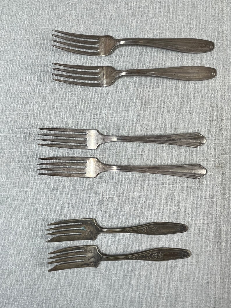 May include: A collection of six silver-toned metal forks arranged in three pairs. The top two forks have smooth handles, the middle two feature a fluted design, and the bottom two display an intricate embossed pattern on the handles.