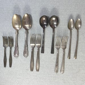 May include: A collection of twelve silver-toned metal utensils arranged in rows, including four forks with ornate handles, two large spoons with decorative stems, two round spoons with plain handles, two smaller forks, and two slender spoons.
