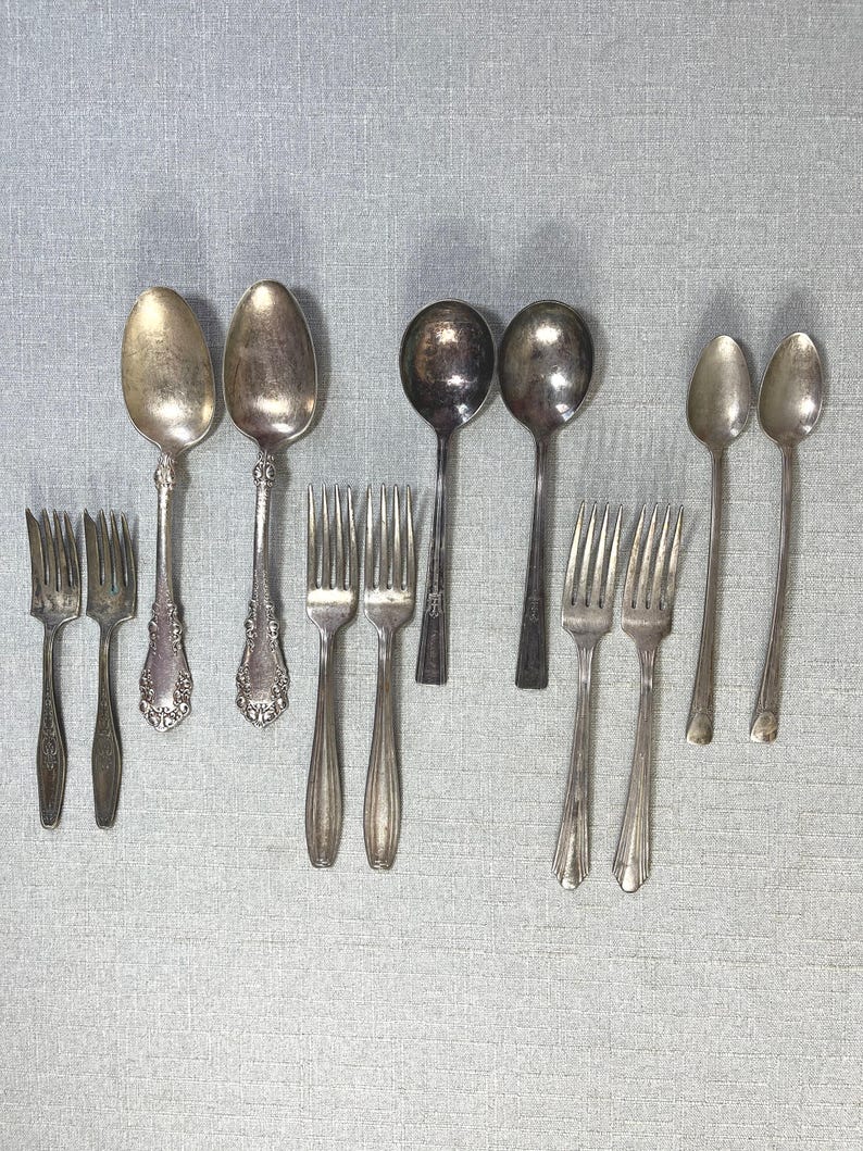 May include: A collection of twelve silver-toned metal utensils arranged in rows, including four forks with ornate handles, two large spoons with decorative stems, two round spoons with plain handles, two smaller forks, and two slender spoons.