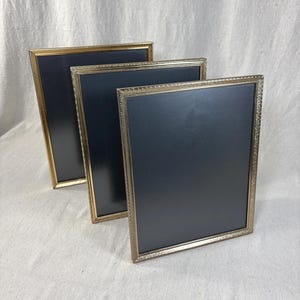 May include: Three rectangular picture frames arranged in a staggered row. Each frame features a metallic gold finish with a textured, beaded edge detail and contains a solid black insert. The frames are positioned upright in a series.