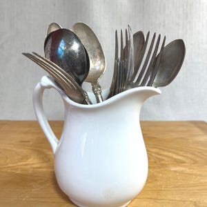 May include: A white ceramic jug containing a collection of assorted silver-toned metal spoons and forks. The jug features a curved handle and a pouring spout, while the cutlery displays various handle designs and metallic finishes.