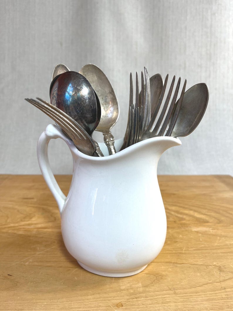 May include: A white ceramic jug containing a collection of assorted silver-toned metal spoons and forks. The jug features a curved handle and a pouring spout, while the cutlery displays various handle designs and metallic finishes.