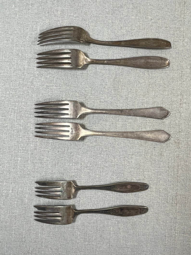May include: Six silver-toned metal forks arranged in three pairs. The top two forks have rounded handles, the middle two feature pointed, angular handle ends, and the bottom two display ornate, decorative detailing on the handles with four tines each.