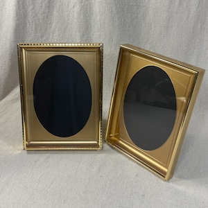 May include: Two rectangular gold-toned picture frames with oval-shaped openings. Each frame features a smooth, metallic finish and a beaded edge detail around the inner border, designed to hold a photograph within the central oval cutout.