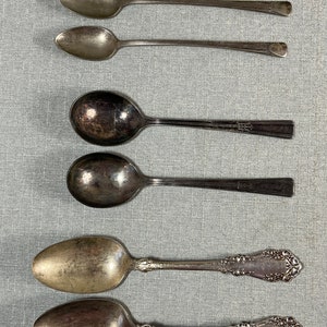 May include: A collection of six silver-toned metal spoons arranged in three pairs. The top pair consists of two slender, plain-handled spoons. The middle pair features two spoons with wide, rounded bowls and simple, flat handles. The bottom pair has ornate, embossed floral detailing on the handles.