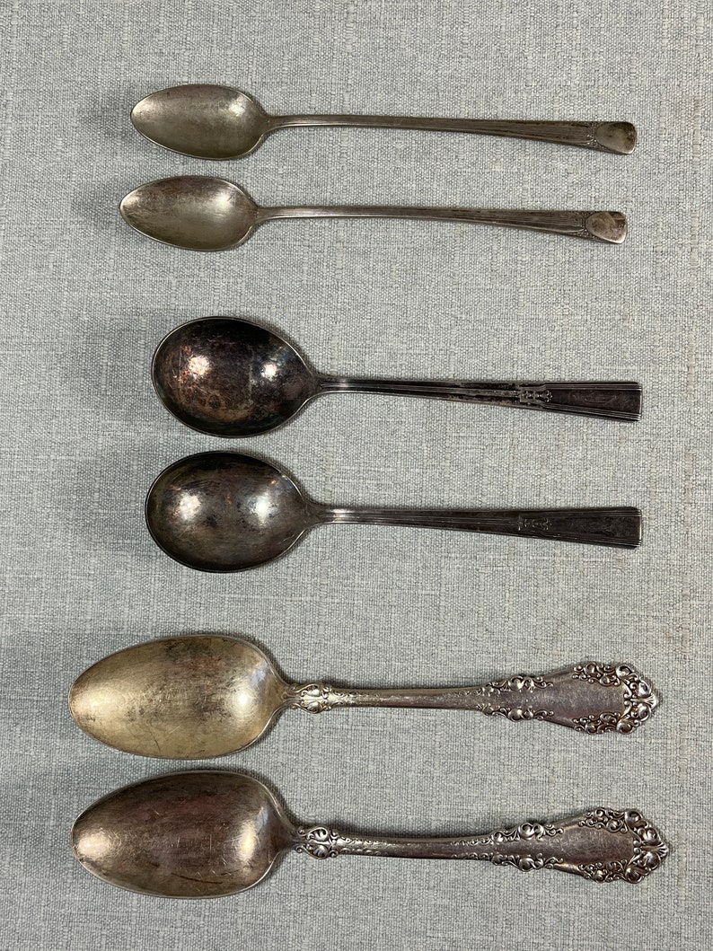 May include: A collection of six silver-toned metal spoons arranged in three pairs. The top pair consists of two slender, plain-handled spoons. The middle pair features two spoons with wide, rounded bowls and simple, flat handles. The bottom pair has ornate, embossed floral detailing on the handles.