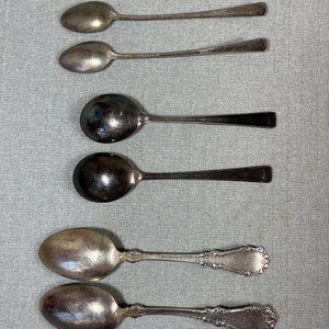 May include: A collection of six silver-toned metal spoons arranged in three pairs. The top two are slender, the middle two have wide round bowls, and the bottom two feature ornate, embossed scrollwork patterns on their handles.