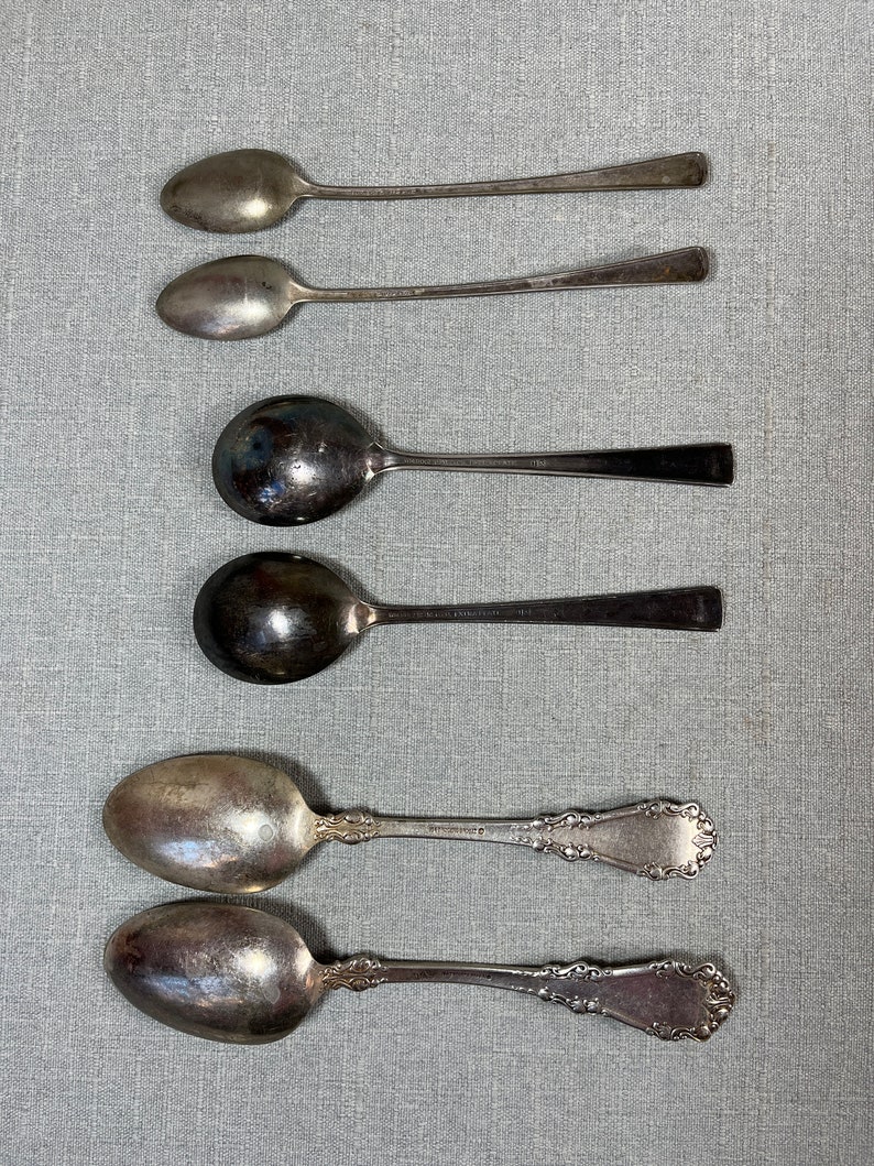 May include: A collection of six silver-toned metal spoons arranged in three pairs. The top two are slender, the middle two have wide round bowls, and the bottom two feature ornate, embossed scrollwork patterns on their handles.