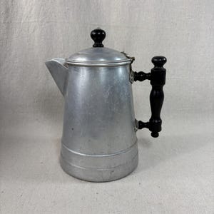 May include: Vintage aluminum stovetop coffee pot with a black wooden handle and lid knob. The pot has a spout for pouring and a hinged lid. The metal has a brushed finish, and the handle is curved.