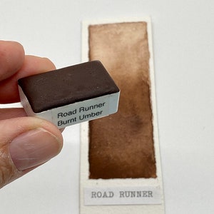 May include: A watercolor paint pan labeled "Road Runner Burnt Umber" with a brown watercolor swatch on a white card labeled "Road Runner".