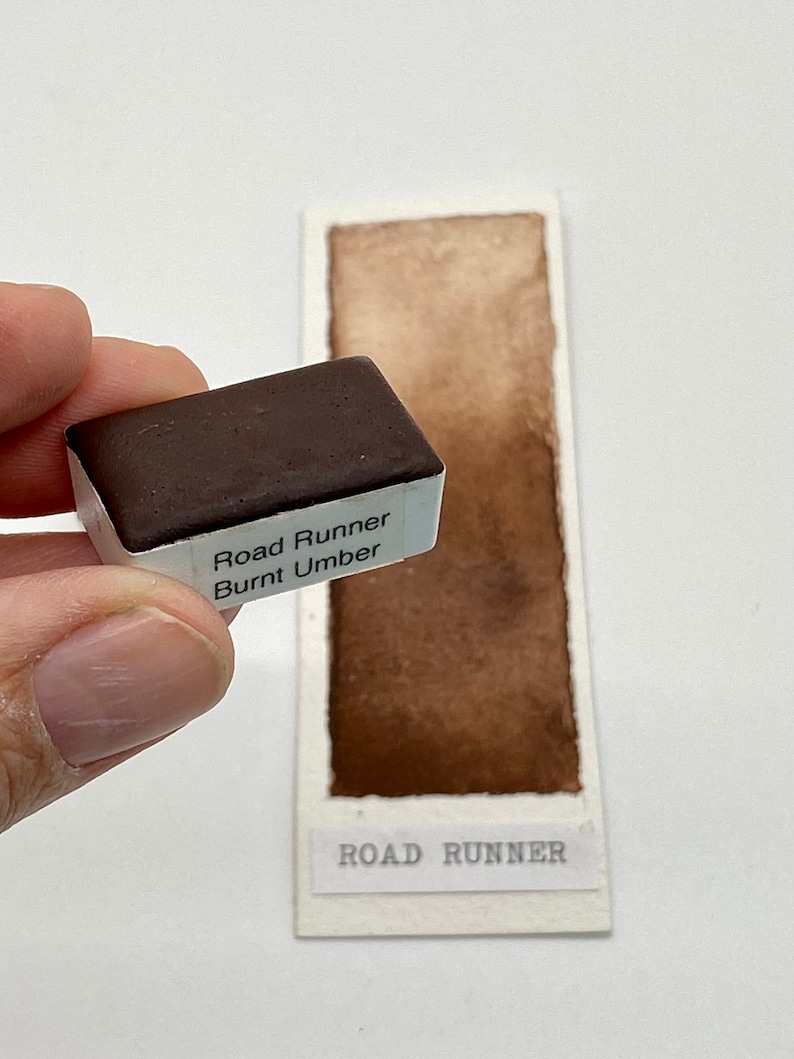 May include: A watercolor paint pan labeled "Road Runner Burnt Umber" with a brown watercolor swatch on a white card labeled "Road Runner".