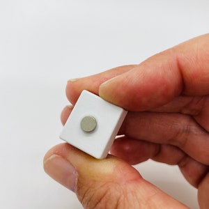 May include: A small white square magnet with a round silver center.