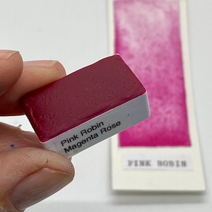 Puede incluir: Primer plano de una pastilla de pintura acuarela rosa etiquetada como "Pink Robin Magenta Rose".