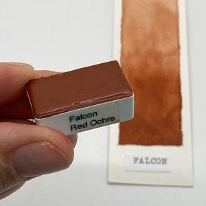 Könnte beinhalten: Eine kleine, quadratische, weiße Schale mit roter Ocker-Aquarellfarbe. Die Schale ist mit "Falcon Red Ochre" beschriftet. Ein Muster der Farbe ist auf einer weißen Karte dargestellt.