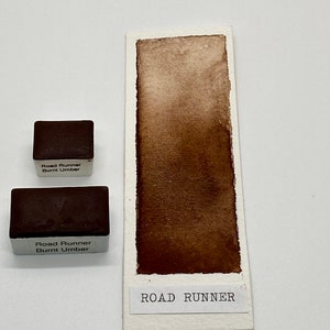 May include: Two watercolor paint pans labeled "Road Runner Burnt Umber" with a brown watercolor swatch labeled "ROAD RUNNER" on a white background.