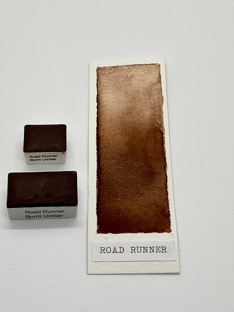 May include: Two watercolor paint pans labeled "Road Runner Burnt Umber" with a brown watercolor swatch labeled "ROAD RUNNER" on a white background.