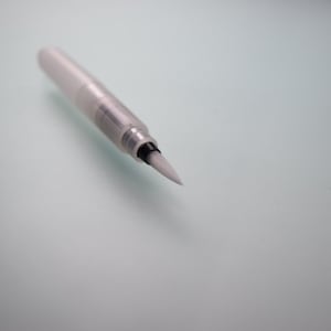 May include: A clear, water brush pen with a white, angled brush tip. The pen's barrel is transparent, revealing the internal components. The brush tip is made of synthetic bristles. The pen is designed for watercolor painting and calligraphy.