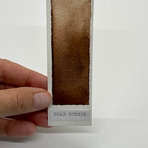 May include: A brown watercolor swatch on a white card with the text "ROAD RUNNER" printed on the bottom.