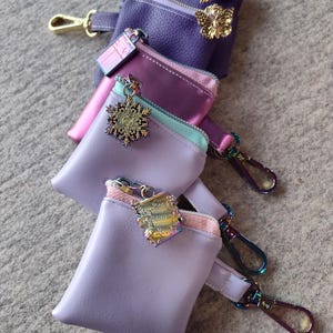 May include: A stack of small, zippered pouches in shades of purple, pink, and lavender. Each pouch has a metal charm and a clip. The top pouch has a gold butterfly charm. The pouches are made of a smooth material.