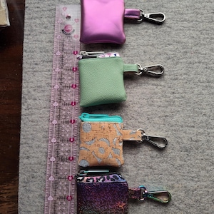 Puede incluir: Cuatro pequeños monederos cuadrados con cierre de cremallera y cierres metálicos. Los monederos son de color rosa metalizado, verde, corcho con detalles plateados y un estampado arcoíris. Una regla rosa está junto a los monederos.