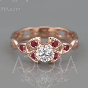 Puede incluir: Un anillo de compromiso de oro rosa con un diamante redondo central y cinco piedras de acento de rubí. El anillo presenta un diseño delicado e intrincado con un motivo de nudo celta.