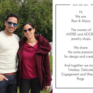 May include: Two people, a man and a woman, stand in front of a green bush. The man is wearing a white sweater, blue jeans, and sunglasses. The woman is wearing a burgundy sweater, a long burgundy coat, and sunglasses. The text on the sign behind them reads: "WHO ARE WE? Hi, We are Reut & Maoz. The owners of AVERIE and ADORA jewelry shops. We share the same passion for design and creation. And together we make Timeless, Delicate Engagement and Wedding Rings."