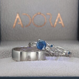 Op de afbeelding: Drie zilveren ringen, waaronder een brede bandring en twee met blauwe edelstenen, gepresenteerd in een grijze doos. De doos is voorzien van het woord "ADORA" in goud. Geschikt voor bruiloft of verloving.
