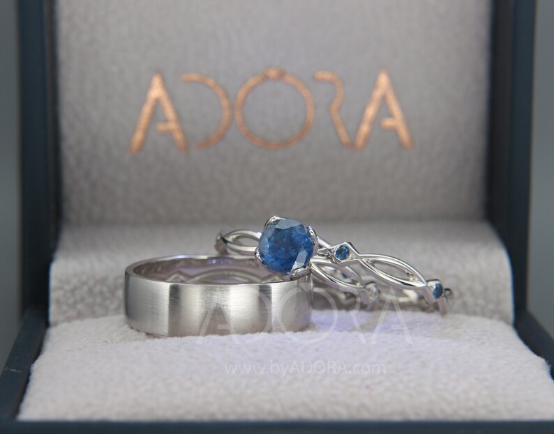 Op de afbeelding: Drie zilveren ringen, waaronder een brede bandring en twee met blauwe edelstenen, gepresenteerd in een grijze doos. De doos is voorzien van het woord "ADORA" in goud. Geschikt voor bruiloft of verloving.