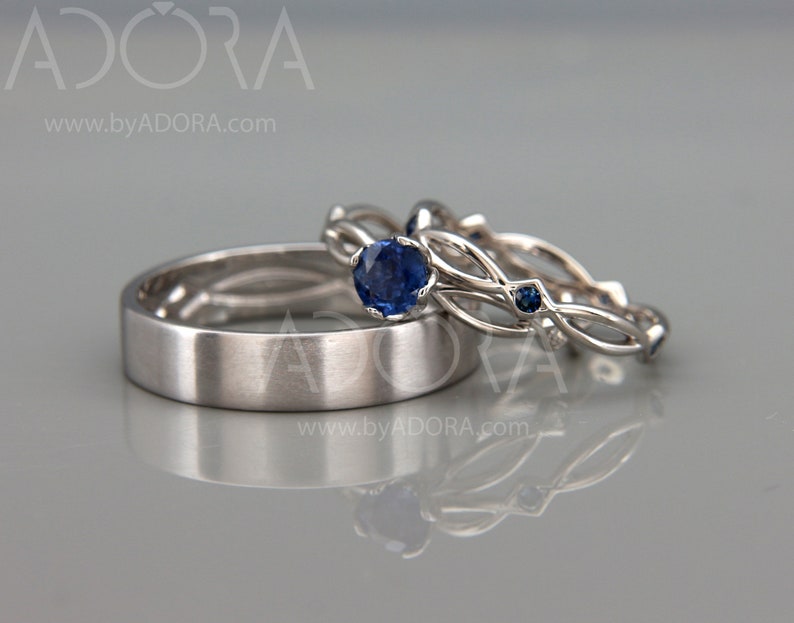 Op de afbeelding: Drie zilveren trouwringen. E&eacute;n is een eenvoudige band, de andere twee hebben een ingewikkeld ontwerp met lussen en kleine, ronde, blauwe edelstenen. De ringen worden gepresenteerd op een reflecterend oppervlak.