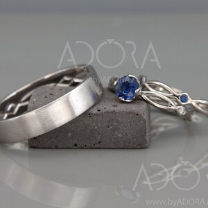 Op de afbeelding: Drie zilveren ringen worden getoond. E&eacute;n ring heeft een geometrisch ontwerp, terwijl de andere twee blauwe edelstenen hebben. E&eacute;n ring heeft een grote, ronde blauwe edelsteen, de andere kleinere blauwe edelstenen. Ze liggen op een grijze ondergrond.