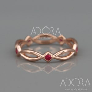 Könnte beinhalten: Roségoldener Ring mit Unendlichkeitsdesign, mit kleinen, leuchtend roten Edelsteinen. Das Band des Rings hat ein kompliziertes, ineinander verwobenes Muster, mit einem Edelstein in regelmäßigen Abständen. Der Ring wird vor einem neutralen grauen Hintergrund präsentiert.