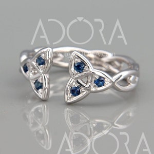 Könnte beinhalten: Ein silberner Ring mit einem keltischen Knotendesign und blauen Edelsteinen. Der Ring ist an einer Seite offen und hat ein filigranes, kompliziertes Design.
