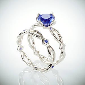 Op de afbeelding: Een set van twee zilveren ringen. E&eacute;n ring heeft een grote, ronde, blauwe edelsteen in een bloemvormige zetting. De tweede ring heeft een gevlochten ontwerp met kleine blauwe edelstenen en een diamantaccent. De ringen worden op een witte achtergrond getoond.