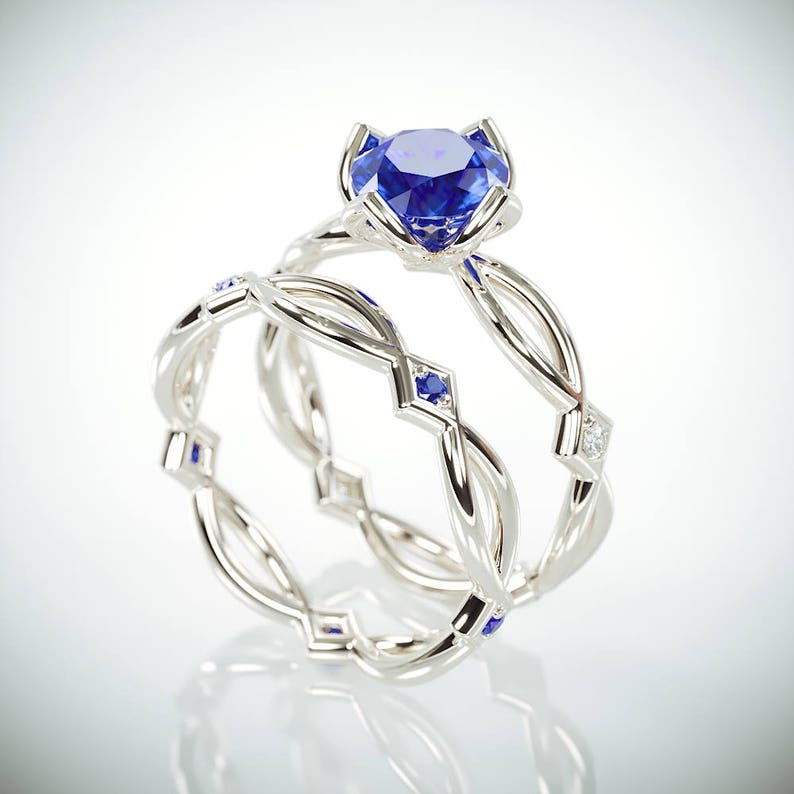 Op de afbeelding: Een set van twee zilveren ringen. E&eacute;n ring heeft een grote, ronde, blauwe edelsteen in een bloemvormige zetting. De tweede ring heeft een gevlochten ontwerp met kleine blauwe edelstenen en een diamantaccent. De ringen worden op een witte achtergrond getoond.