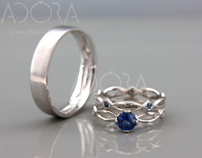Op de afbeelding: Twee zilveren ringen. E&eacute;n is een eenvoudige band met een geborstelde afwerking. De andere is een set van twee ringen met een verweven ontwerp, met een grote, ronde, donkerblauwe edelsteen en kleinere blauwe accentstenen.