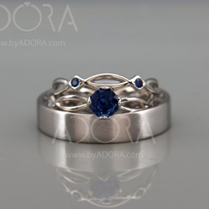 Op de afbeelding: Drie zilveren ringen met blauwe edelstenen. De middelste ring heeft een grote, ronde saffier, terwijl de andere ringen kleinere blauwe stenen hebben. De ringen staan tegen een lichtgrijze achtergrond.