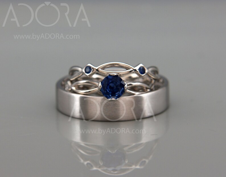 Op de afbeelding: Drie zilveren ringen met blauwe edelstenen. De middelste ring heeft een grote, ronde saffier, terwijl de andere ringen kleinere blauwe stenen hebben. De ringen staan tegen een lichtgrijze achtergrond.