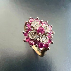 Puede incluir: Un anillo dorado con un grupo de gemas redondas de color rosa intenso y pequeñas piedras transparentes. El diseño del anillo se asemeja a un arreglo floral, con las gemas muy juntas. La banda es de oro pulido simple.