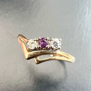 May include: A gold-toned ring featuring three stones: a central purple gemstone flanked by two clear, round-cut stones. The ring has a unique, asymmetrical design with a V-shaped element and a curved band. The gemstones are set in a prong setting.