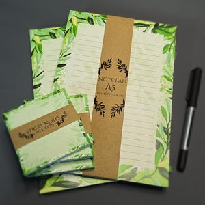 Könnte beinhalten: Ein Schreibwarenset mit grünem Blattmuster, bestehend aus einem A5-Notizblock und Haftnotizen. Der Notizblock hat linierte Seiten und wird von einem braunen Band zusammengehalten. Die Haftnotizen sind mit einem braunen Papierband gebündelt. Ein schwarzer Stift liegt daneben.