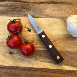 Op de afbeelding: Een roestvrijstalen schilmes met een donkerhouten handvat en messing klinknagels ligt op een houten snijplank naast een tros rode tomaten en een bol knoflook. Op het mes staat de tekst "Carley Clopton".