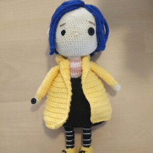May include: Crocheted doll featuring a character with blue hair, a yellow coat, black skirt, and yellow shoes. The doll has a white face with black button eyes and a striped shirt. The doll is handmade and appears to be a character from a movie.