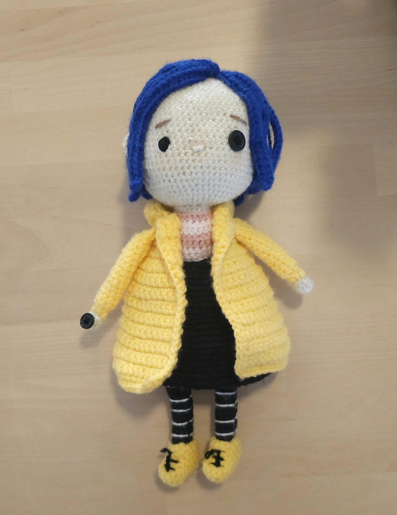 May include: Crocheted doll featuring a character with blue hair, a yellow coat, black skirt, and yellow shoes. The doll has a white face with black button eyes and a striped shirt. The doll is handmade and appears to be a character from a movie.