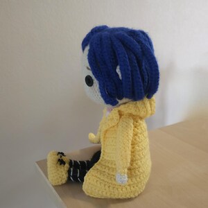 May include: Crocheted doll featuring a character with blue hair, a white face, and a yellow hooded jacket. The doll is seated, with black and white striped pants and black shoes. The doll is handmade and appears to be a character from a movie.